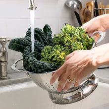 Stainless steel colander under a faucet, filled with leafy greens for draining