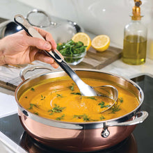 Stainless steel ladle held over soup pot with herbs and citrus on counter