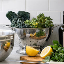 Stainless steel colander with perforated sides, holding greens and lemon wedges