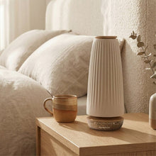 Ceramic diffuser with warm glow on a bedside table near a mug and white linens