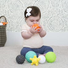 Baby holding an orange textured ball, with teal, gray, white, green, and yellow balls nearby