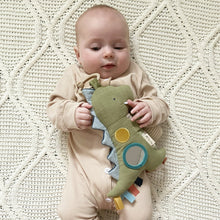 Olive green dino car seat toy held by baby, with teal ring and ribbon tails