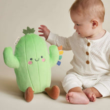 Green cactus plush toy with smiling face, small fabric ribbons, held by a baby