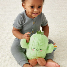 Green cactus plush toy with smiling face and multicolor ribbon arm, held by a toddler