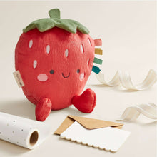 Plush strawberry toy with green leaf, red body, smiling face, and white ribbon strap