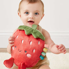 Plush strawberry toy in baby arms; red body, green leaf, stitched smile, ribbon tags