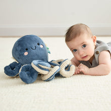 Blue plush octopus with smiling face and ribbon accents lying beside a baby.
