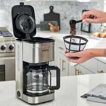 10-cup drip coffee maker with glass carafe and filter, hands filling grounds