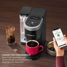 Coffee maker brewing into red mug; hand holds phone while clear carafe sits beside