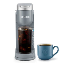 Gray iced coffee maker with blue mug beside a clear cup of iced coffee.