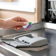 Green, purple and white dishwasher detergent pac held above a dishwasher dispenser