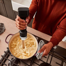 Cordless hand blender held over a skillet, blending attachment submerged