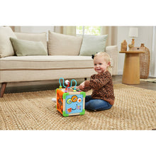 Wooden activity cube toy with handle bars and number panels on a rug, with toddler nearby