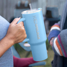 Light blue tumbler with clear straw, etched vertical logo, held in a hand.