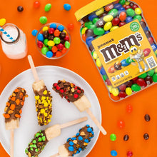 Peanut M&M's candy jar with yellow lid, surrounded by assorted chocolate candy pieces