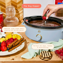 Light blue slow cooker with black lid and fruit-dipping hand over open pot
