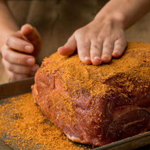 Hands applying orange-brown spice rub to raw pork roast on board.