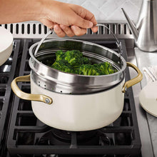 Cream ceramic steamer pot with gold handles on a stove with broccoli