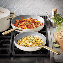 Cream ceramic sauté pan and pot with gold handles on a stove with food