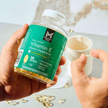 Hands holding Vitamin E softgel bottle with clear container of golden capsules