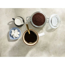 Glass container of dark coffee grounds with lid, scoop, and cup on tray
