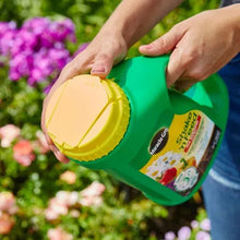 Miracle-Gro Shake 'n Feed jug with yellow cap, held by hands over garden plants