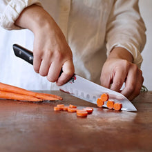 Hands slicing with Miyabi Santoku knife; dark handle and scalloped blade