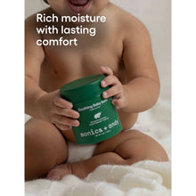 Baby holding dark green soothing baby balm jar on a soft white blanket