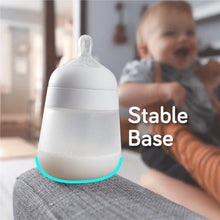 White silicone baby bottle with stable base, next to a drinking baby