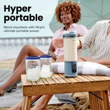 Portable blender with clear jar and blue base on a table beside a seated woman