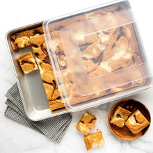 Aluminum roasting pan with clear lid, containing baked bars and scattered pieces