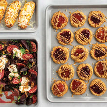 Two non-stick baking sheets with baked cookies on one and roasted food on the other