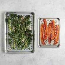 Two perforated metal trays holding greens and cooked chicken on hole patterns