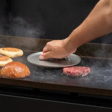 Grill press being lowered by a hand onto food on the ridged cooking surface