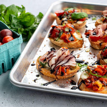 Rectangular aluminum sheet pan with bruschetta-style toasts and sliced toppings
