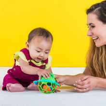 Infant playing with green frog jingle rattle, reaching toward the colorful handles