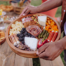 Round acacia wood serving tray with glass insert, filled with assorted snacks and hands