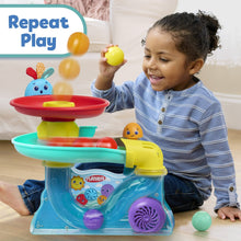 Busy ball popper toy with red top disc and yellow launcher, toddler playing beside it.