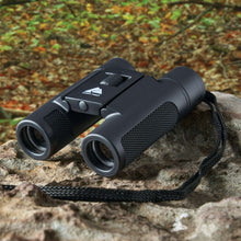 Black pocket binoculars with neck strap resting on a rock outdoors.