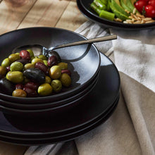 Dark dinner plate and bowl with serving spoon, holding olives in foreground