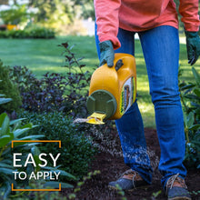 Weed preventer bottle being poured onto garden soil beside shrubs.