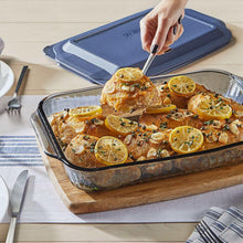 Tinted glass baking dish with blue lid beside another dish; hand garnishing roast