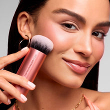 Makeup blending brush held near cheek, rose-gold handle with white bristles.