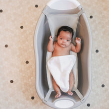 Grey plastic infant bath tub with soft mesh sling, holding a baby for bathing