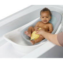 White baby bathtub with infant reclining in Harbor Mist insert, caregiver hand visible