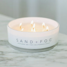 White scented candle in glass jar with lit wicks and Sand + Fog logo