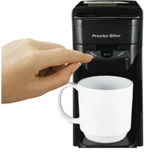 Black single-serve coffee maker with white mug and hand at controls