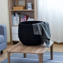 Black knit storage basket with a folded dark throw blanket draped over side