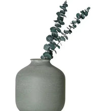 Green metal tabletop vase with angled eucalyptus stems, wide body and short neck.