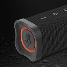 Black rugged wireless speaker with orange-ring top panel on wet textured surface